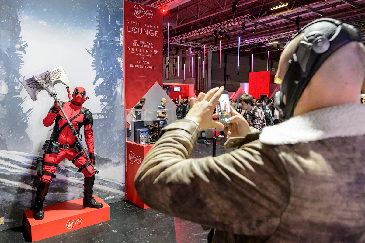 A fan at a MatterXP event capturing a picture of a cosplayer dressed in a red and black superhero outfit, holding an axe in a dynamic pose near a gaming lounge booth.